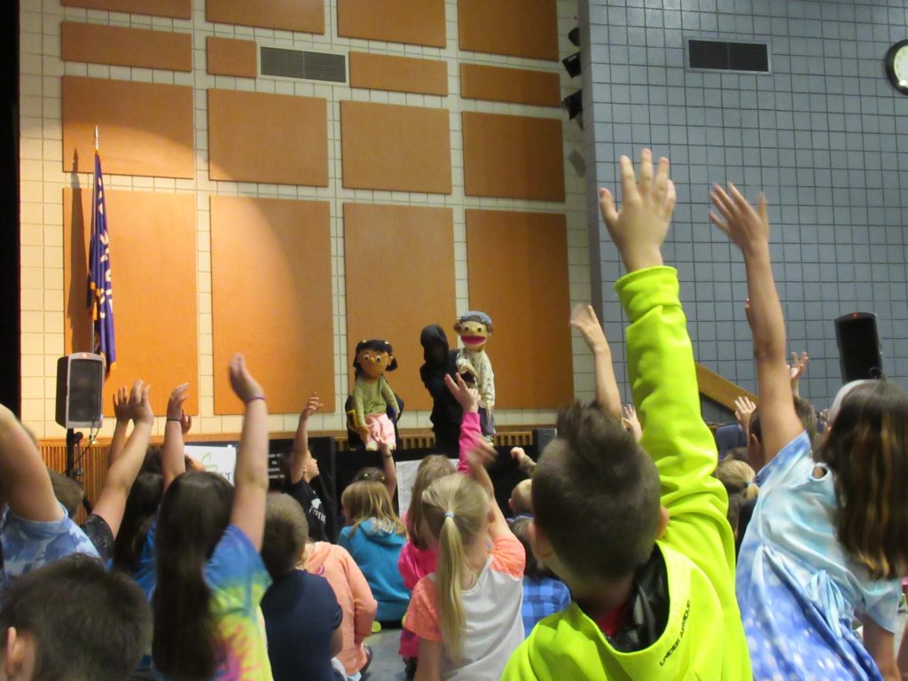Puppets in Education performance image with children's hands raised.