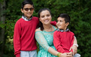 A parent smiles while surrounded by their two children. One child wears dark glasses.